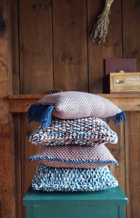 A stack of four Halcyon Yarn's Eastport Pillows, featuring an array of woven designs and colors akin to a knitting project, in blue and earthy tones, is arranged on a green wooden stool against a rustic wooden wall. In the background, a small shelf holds a box labeled 'Lighthouse Crackers' and a dried bunch of herbs.