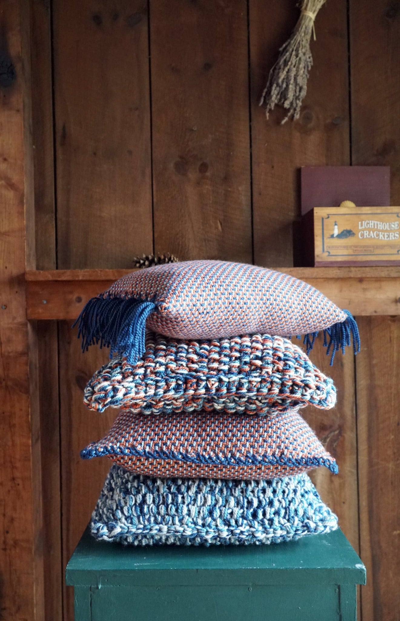 A stack of four Eastport Pillows, crafted using seed stitch with various textures and colors, featuring predominantly blue and orange patterns by Halcyon Yarn, sits atop a small green stool. The background is a rustic wooden wall with a shelf holding a box labeled "Lighthouse Crackers" and a bunch of dried herbs.