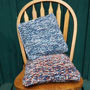 Two Eastport Pillows by Halcyon Yarn are arranged on a wooden chair with a vertical slat back. The top cushion features a blue, white, and gray color pattern, while the bottom cushion displays a blend of blue, brown, and white colors. The chair is set against a green wooden background.