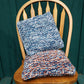 Two Eastport pillows from Halcyon Yarn rest on a wooden chair with a green background. The top cushion is in shades of blue, gray, and white, while the bottom cushion features a mix of blue, white, and orange yarn. The textured pattern adds a cozy and handmade feel.