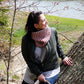 A person with dark hair and a red and white striped Popham Cowl from Halcyon Yarn, crafted from Aran weight yarn, leans against a tree near a river. They are wearing a green jacket and light-colored shirt, smiling and looking to the side. Budding branches are visible in the background.