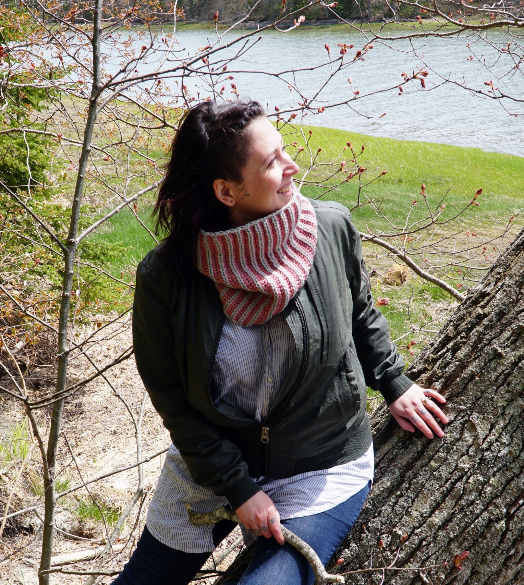 A person with dark hair and a red and white striped Popham Cowl from Halcyon Yarn, crafted from Aran weight yarn, leans against a tree near a river. They are wearing a green jacket and light-colored shirt, smiling and looking to the side. Budding branches are visible in the background.