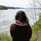 A person with shoulder-length dark hair, wearing a brown jacket and a pink scarf that resembles the Popham Cowl from Halcyon Yarn, stands with their back to the camera, gazing out at a large body of water surrounded by trees. The scene is serene, with muted colors indicating early spring or late fall.