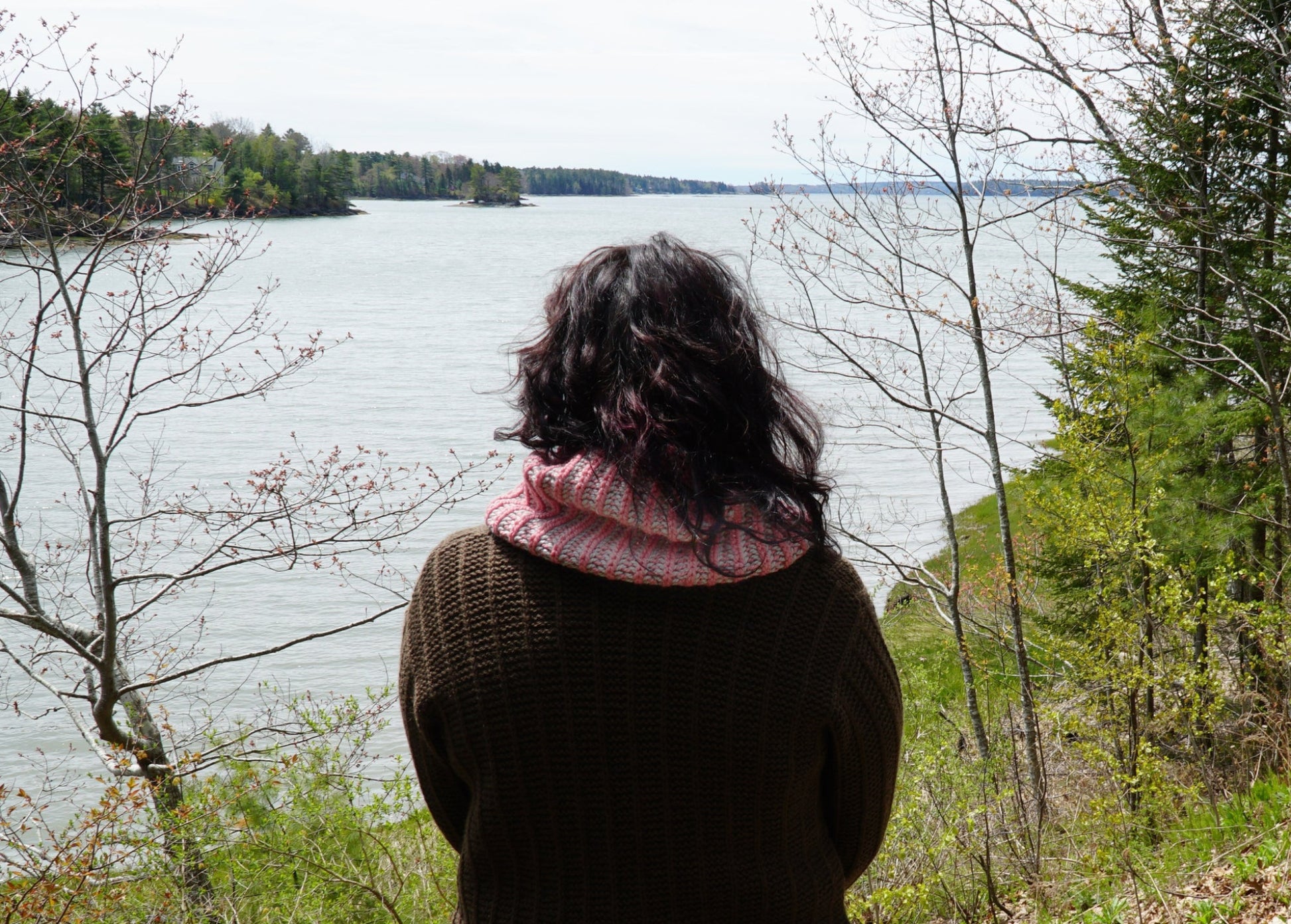 A person with shoulder-length dark hair, wearing a brown jacket and a pink scarf that resembles the Popham Cowl from Halcyon Yarn, stands with their back to the camera, gazing out at a large body of water surrounded by trees. The scene is serene, with muted colors indicating early spring or late fall.