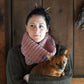 A woman with dark hair tied up, adorned with two knitting needles, wears a Popham Cowl and a large sweater made from Aran weight yarn by Halcyon Yarn. Holding a small brown dog in her arms, she stands against a wooden wall that lends a rustic feel.