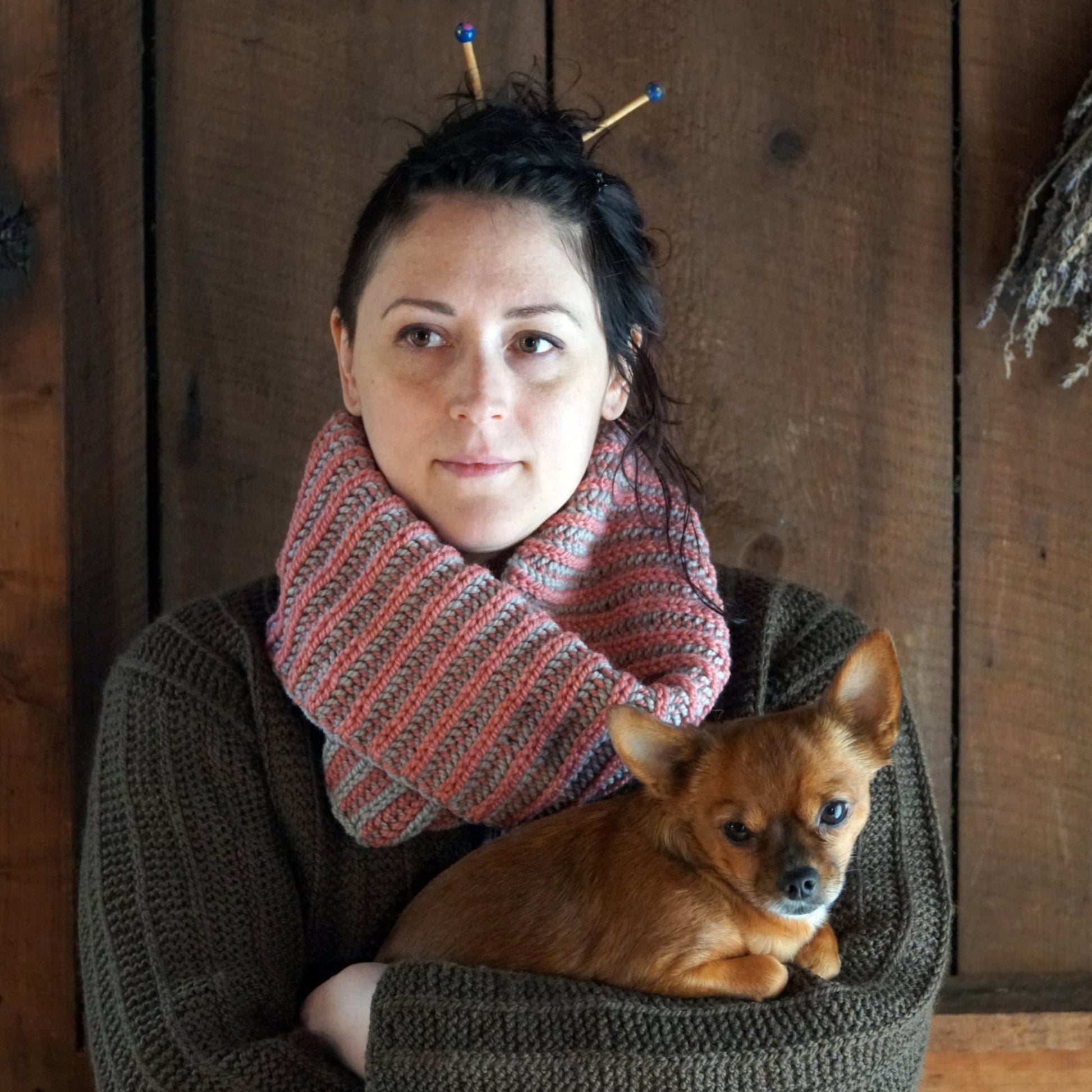 A woman with dark hair tied up, adorned with two knitting needles, wears a Popham Cowl and a large sweater made from Aran weight yarn by Halcyon Yarn. Holding a small brown dog in her arms, she stands against a wooden wall that lends a rustic feel.