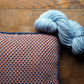 A squishy Halcyon Yarn Vacationland Woven Pillow with a red, white, and blue houndstooth pattern lies on a wooden surface. Resting on the pillow is a skein of light blue Norumbega yarn.