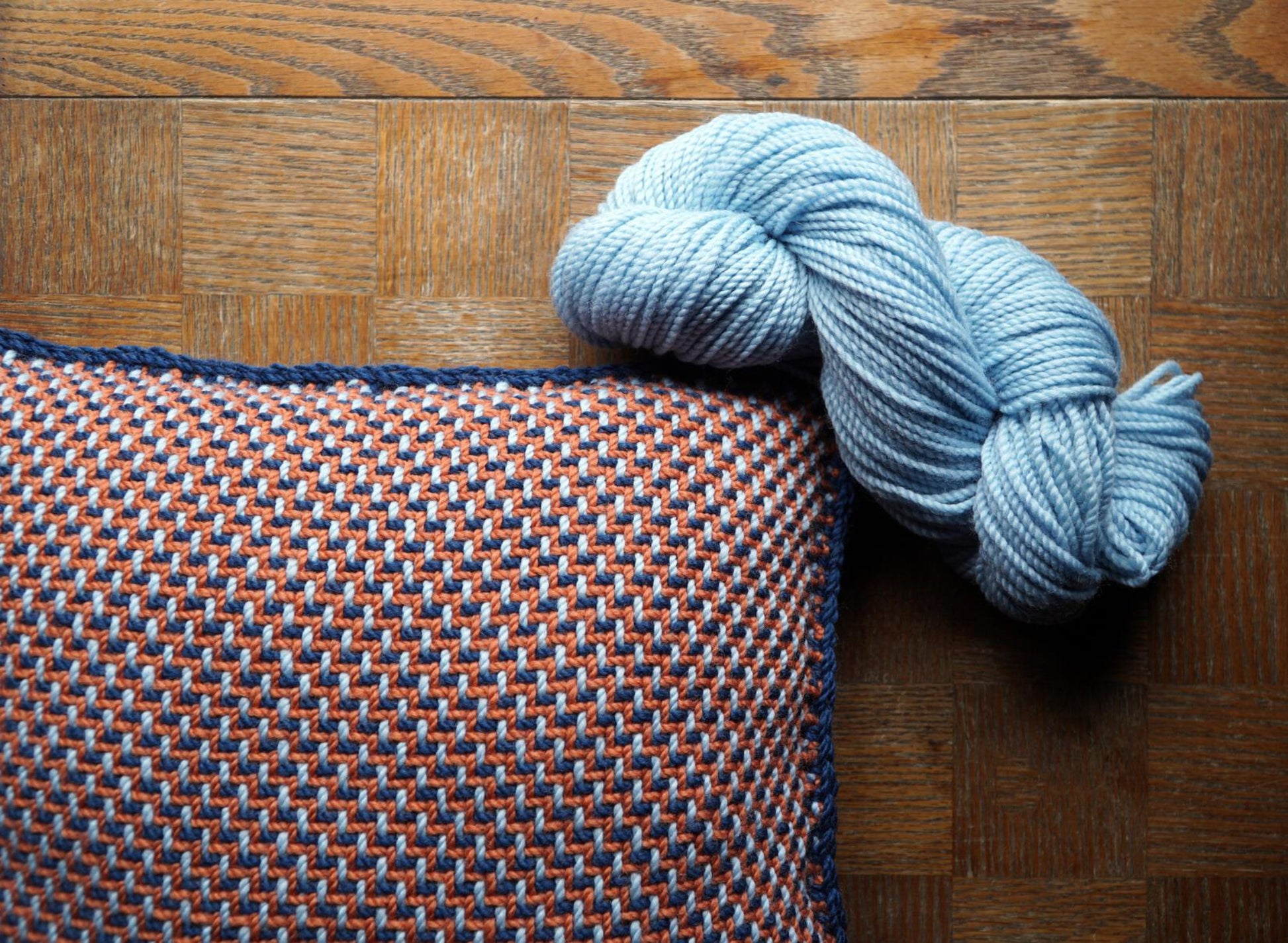 A squishy Halcyon Yarn Vacationland Woven Pillow with a red, white, and blue houndstooth pattern lies on a wooden surface. Resting on the pillow is a skein of light blue Norumbega yarn.