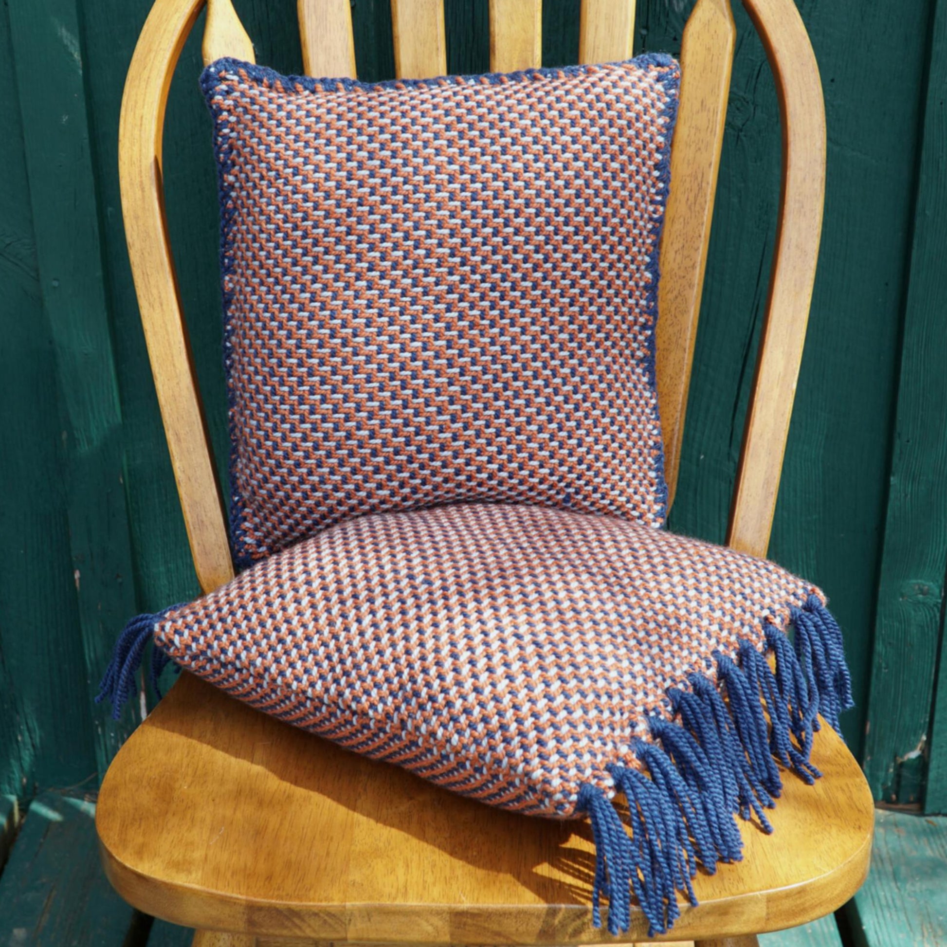 A wooden chair with a green background features two squishy pillows: one propped against the chair back and the other resting on the seat. Both Vacationland Woven Pillows, crafted by Halcyon Yarn, showcase a checkered twill pattern and blue fringes.