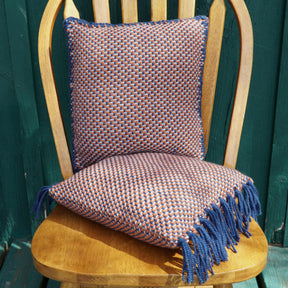 A wooden chair with a square woven cushion from Halcyon Yarn's Vacationland Woven Pillows collection, featuring a blue and tan checkered, color-and-weave twill pattern on the seat, and a matching tassel-accented cushion draped on the backrest. The chair is set against a dark green wall.