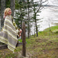 A person wrapped in a Chickadee Woven Blanket from Halcyon Yarn sits on a wooden fence, smiling, with a scenic view of trees, grass, and a body of water in the background. The day appears overcast and serene, suggesting a peaceful outdoor setting.