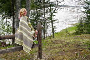 A person wrapped in a Chickadee Woven Blanket from Halcyon Yarn sits on a wooden fence, smiling, with a scenic view of trees, grass, and a body of water in the background. The day appears overcast and serene, suggesting a peaceful outdoor setting.