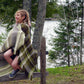 A person with shoulder-length hair sits on a wooden fence, wrapped in a large, green and brown Chickadee Woven Blanket by Halcyon Yarn. They are smiling and looking away from the camera. Behind them, there is a serene lake surrounded by trees and greenery.