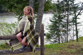 A person with shoulder-length hair sits on a wooden fence, wrapped in a large, green and brown Chickadee Woven Blanket by Halcyon Yarn. They are smiling and looking away from the camera. Behind them, there is a serene lake surrounded by trees and greenery.