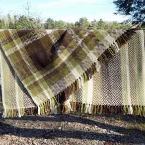 The Chickadee Woven Blanket by Halcyon Yarn, featuring green, brown, and cream six-point herringbone twill, is draped over a line outdoors. Its advanced beginner weaving techniques and natural colors harmonize with the trees and blue sky in the background, creating a rustic and cozy ambiance.