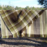 A thick, woven Chickadee blanket from Halcyon Yarn with a herringbone pattern hangs outdoors on a line. The blanket, known as the Chickadee Woven Blanket, download, features earthy tones of green, brown, beige, and white with fringes along the edges. Trees and sky are visible in the background.