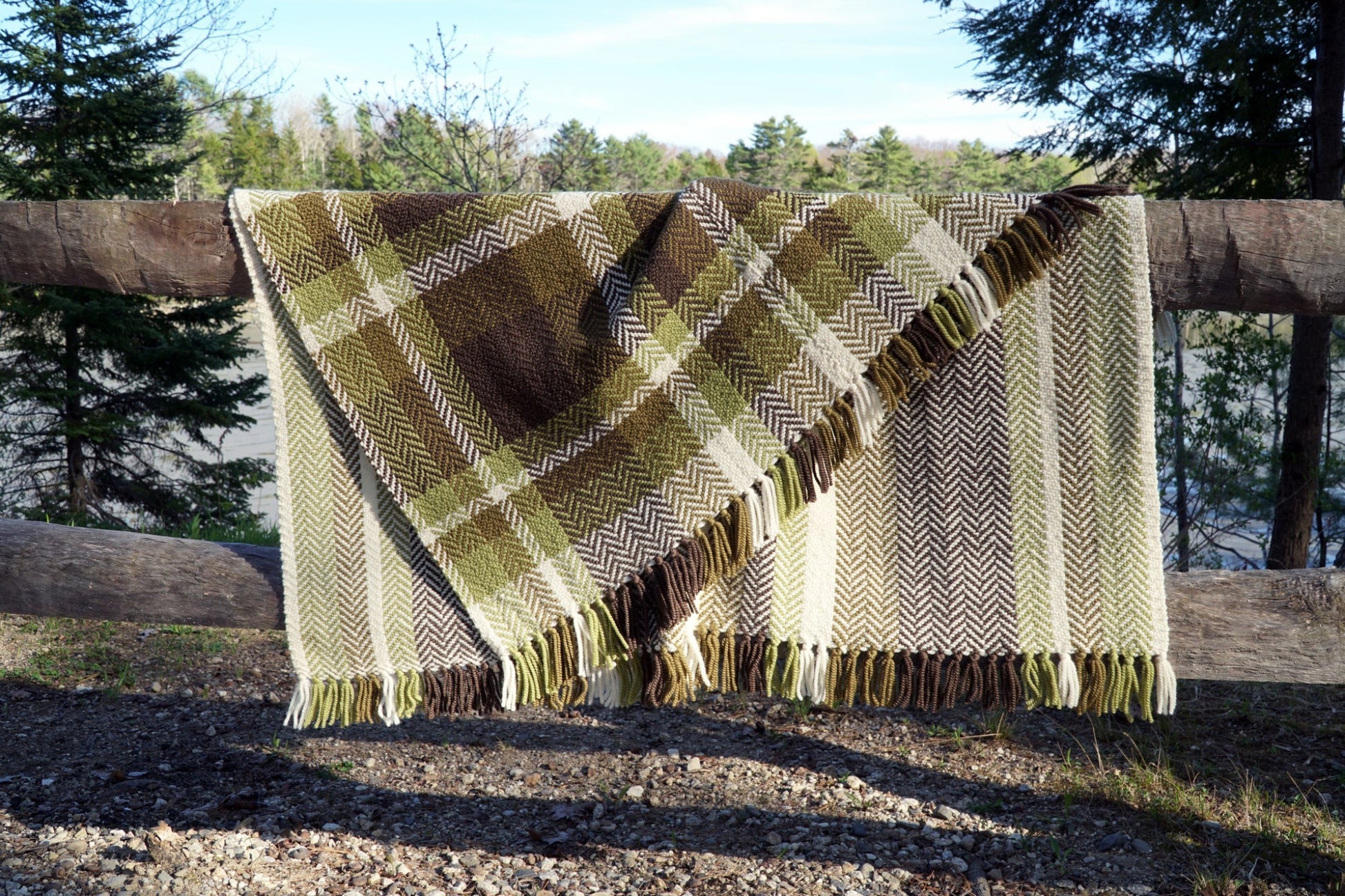 A Chickadee Woven Blanket from Halcyon Yarn, featuring green, brown, and cream colors, is draped over a wooden fence. Designed with varied patterns and fringed edges, this six-point herringbone twill adds charm to the natural outdoor setting with trees and a lake in the background.