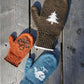 Three Pemaquid Mittens from Halcyon Yarn are laid out on a wooden surface: a child's orange mitten with a brown tree design, an adult's brown mitten with a white tree design, and an adult's blue mitten with a white snowflake design, showcasing our family-sized customizable mittens.