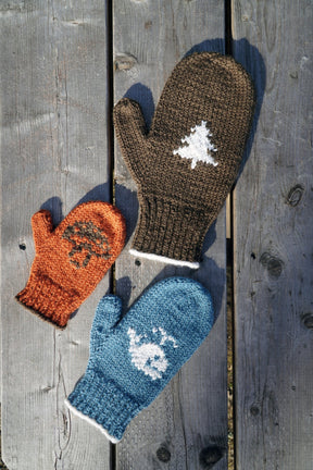 Three Pemaquid Mittens from Halcyon Yarn are laid out on a wooden surface: a child's orange mitten with a brown tree design, an adult's brown mitten with a white tree design, and an adult's blue mitten with a white snowflake design, showcasing our family-sized customizable mittens.