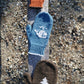 Three Pemaquid Mittens displayed on a wooden plank: the top one is orange with a sun-like pattern, the middle one is blue with a white whale design, and the bottom one is brown with a white tree motif. These family-sized mittens by Halcyon Yarn showcase intricate knitting patterns against a background of gravelly ground.