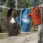 Three vibrant, family-sized Halcyon Yarn Pemaquid Mittens hang on a clothesline with clothespins in an outdoor setting. The brown mitten has a white tree pattern, the blue mitten has a white whale pattern, and the orange mitten features a brown mushroom knitting pattern.