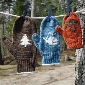 Three vibrant, family-sized Halcyon Yarn Pemaquid Mittens hang on a clothesline with clothespins in an outdoor setting. The brown mitten has a white tree pattern, the blue mitten has a white whale pattern, and the orange mitten features a brown mushroom knitting pattern.