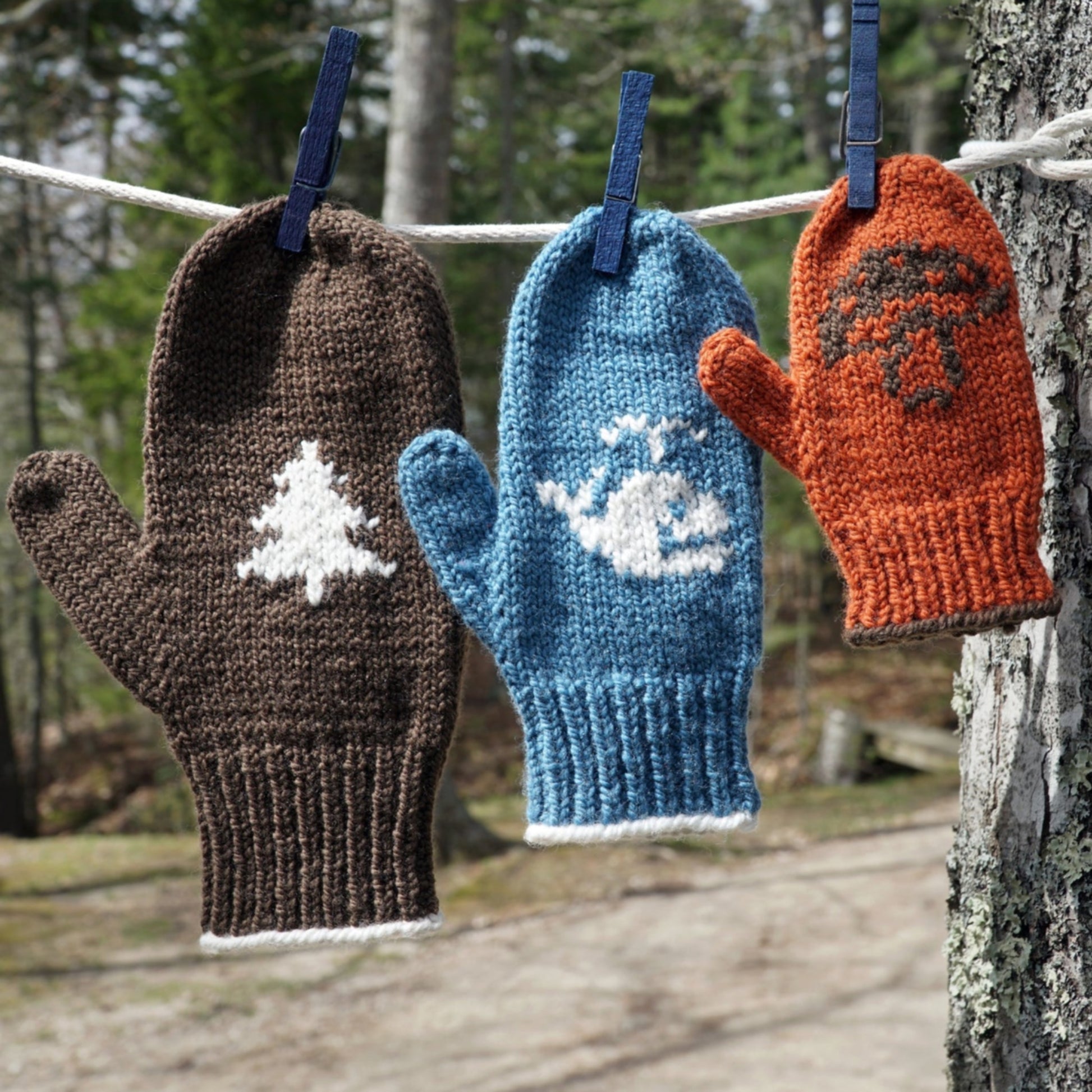 Three knitted, customizable Pemaquid Mittens offered by Halcyon Yarn hang on a clothesline in a wooded area. The brown mitten has a white tree design, the blue mitten features a white whale, and the orange mitten shows a brown mushroom. Each is clipped with a blue clothespin.