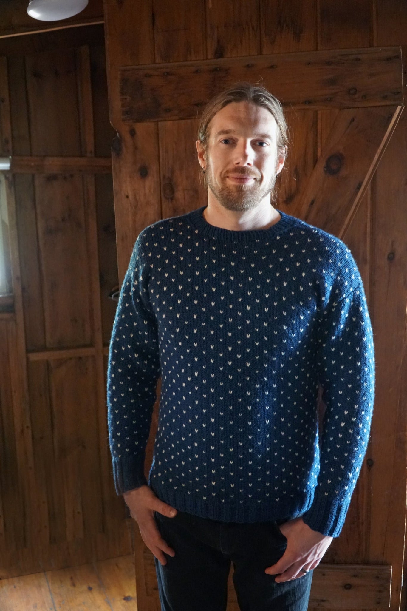 A person with shoulder-length blond hair and a beard is standing in a wooden room. They are wearing the blue Halcyon Yarn Lubec Pullover, which features a charming white snowflake pattern—ideal for beginners learning colorwork. The rustic wooden walls and minimal lighting of the room create a cozy atmosphere.