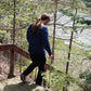 A person with long hair and a blue patterned Halcyon Yarn Lubec Pullover walks down wooden steps surrounded by trees and foliage on a sunny day. A body of water is visible through the trees in the background.