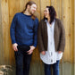 A man and a woman stand side by side against a wooden wall, laughing and looking at each other. The man, with long hair, is dressed in a blue Halcyon Yarn Lubec Pullover paired with jeans and sneakers. The woman has dark hair and wears a brown cardigan over a long shirt, black leggings, and red shoes.