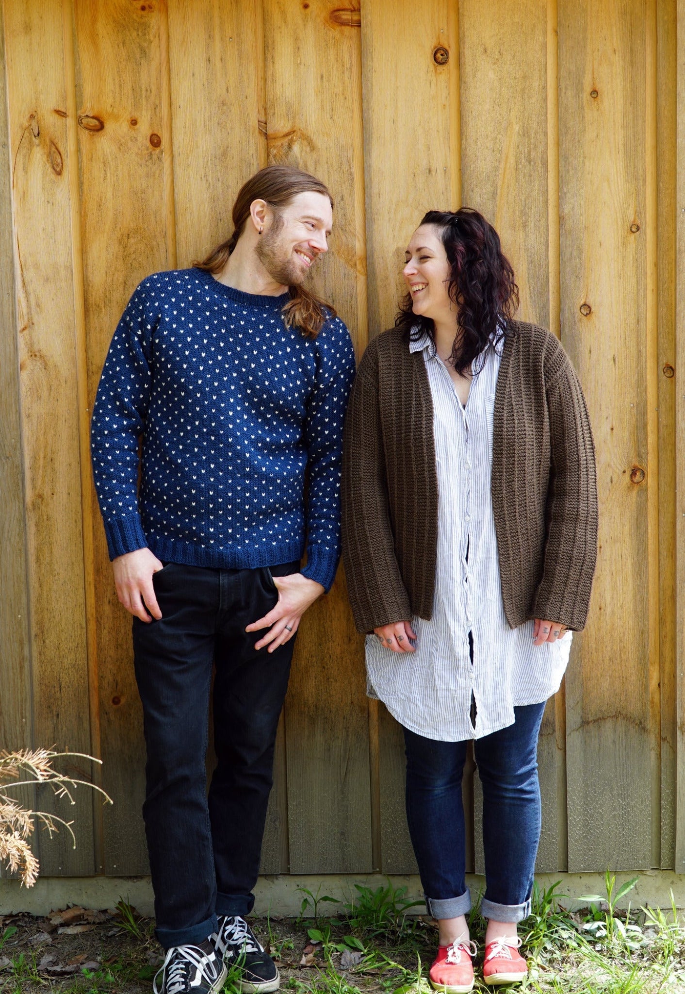 A man and a woman stand side by side against a wooden wall, laughing and looking at each other. The man, with long hair, is dressed in a blue Halcyon Yarn Lubec Pullover paired with jeans and sneakers. The woman has dark hair and wears a brown cardigan over a long shirt, black leggings, and red shoes.