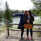 A man and a woman stand near a wooden fence with a scenic lake and trees in the background. The man, wearing the Halcyon Yarn Lubec Pullover, is pointing off into the distance while standing on one leg. The woman, holding a picnic basket, wears a plaid shirt and looks in the same direction.