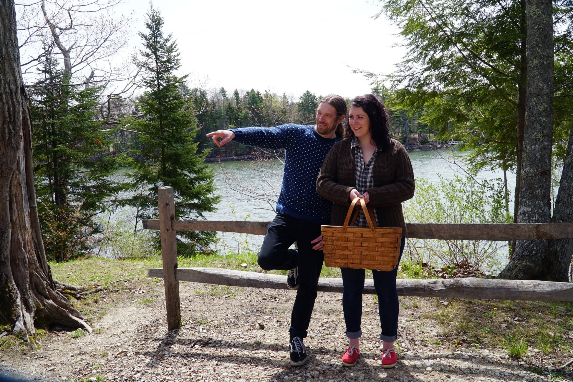 A man and a woman stand near a wooden fence with a scenic lake and trees in the background. The man, wearing the Halcyon Yarn Lubec Pullover, is pointing off into the distance while standing on one leg. The woman, holding a picnic basket, wears a plaid shirt and looks in the same direction.