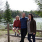 A family of three poses outdoors by a wooden fence with a forested lake in the background. The young girl sits on the fence wearing a red striped sweater styled like the Lubec Pullover from Halcyon Yarn, while the father and mother, both wearing sweaters, stand beside her smiling. Trees and water are visible behind them.