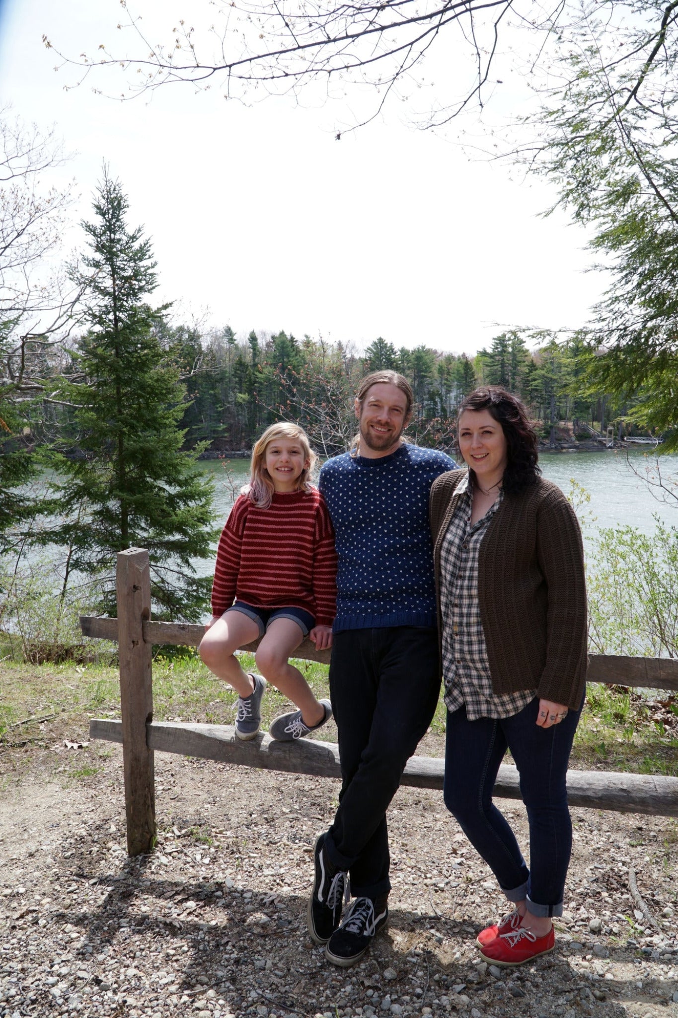 A family of three poses outdoors by a wooden fence with a forested lake in the background. The young girl sits on the fence wearing a red striped sweater styled like the Lubec Pullover from Halcyon Yarn, while the father and mother, both wearing sweaters, stand beside her smiling. Trees and water are visible behind them.