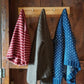 Three knit sweaters, including a drop shoulder crew neck Lubec Pullover from Halcyon Yarn, hang on a wooden wall rack. From left to right, the sweaters are red with white stripes, solid brown, and blue with white polka dots—a perfect beginner's introduction to colorwork against the dark, rustic wooden wall backdrop.