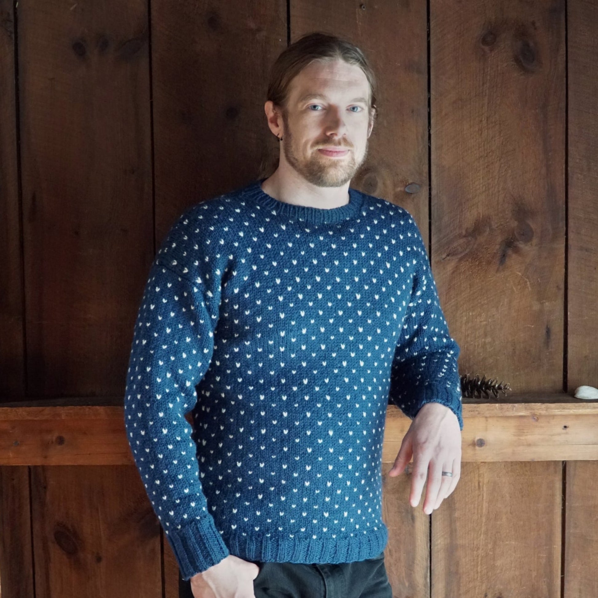 A person with long, light-colored hair and a beard is standing indoors against a wooden wall. They are wearing a blue, patterned Lubec Pullover from Halcyon Yarn and have a relaxed expression, with one hand resting on a wooden surface.