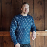 A man with long blonde hair tied back is leaning against a wooden wall. He is wearing the Lubec Pullover, a blue drop shoulder crew neck sweater with a white dotted pattern from Halcyon Yarn, along with black pants. He is looking towards the camera with a relaxed expression. There is a pinecone on a wooden beam behind him.