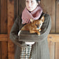 A person dressed in a cozy Rangeley Cardigan by Halcyon Yarn—a knitted brown sweater featuring intricate Garter Rib stitching—stands against a wooden backdrop, clutching a small brown dog. They accessorize with a pink scarf and have knitting needles tucked into their hair.