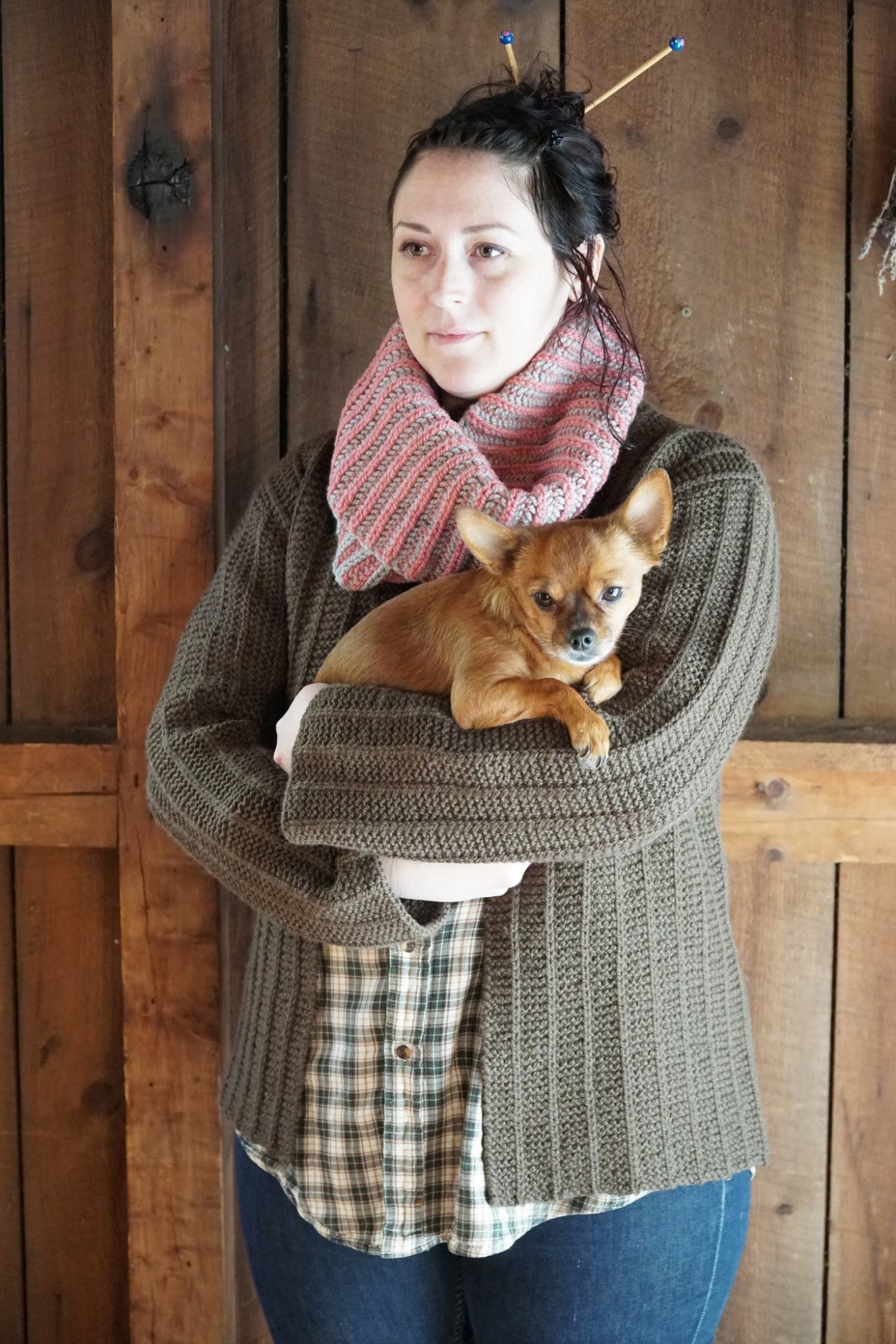 A person dressed in a cozy Rangeley Cardigan by Halcyon Yarn—a knitted brown sweater featuring intricate Garter Rib stitching—stands against a wooden backdrop, clutching a small brown dog. They accessorize with a pink scarf and have knitting needles tucked into their hair.