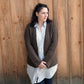 A woman with shoulder-length dark hair stands in front of a wooden wall. She is wearing the Halcyon Yarn Rangeley Cardigan made from Norumbega yarn, displaying the intricate Garter Rib stitch over her long light blue and white striped shirt, paired with dark jeans. She looks into the distance with her hands clasped in front of her.