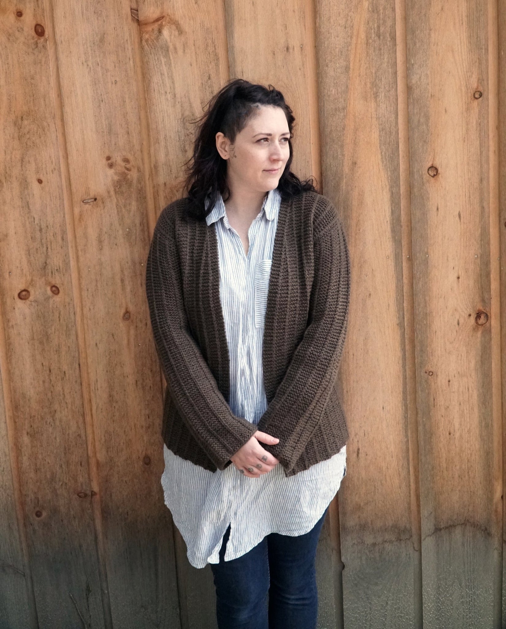 A woman with shoulder-length dark hair stands in front of a wooden wall. She is wearing the Halcyon Yarn Rangeley Cardigan made from Norumbega yarn, displaying the intricate Garter Rib stitch over her long light blue and white striped shirt, paired with dark jeans. She looks into the distance with her hands clasped in front of her.