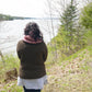 A person with long dark hair, wearing the Rangeley Cardigan by Halcyon Yarn in a Garter Rib stitch along with a white shirt and a red scarf, stands with their back to the camera, overlooking a lake surrounded by trees. The landscape is lush with greenery and the sky is overcast.