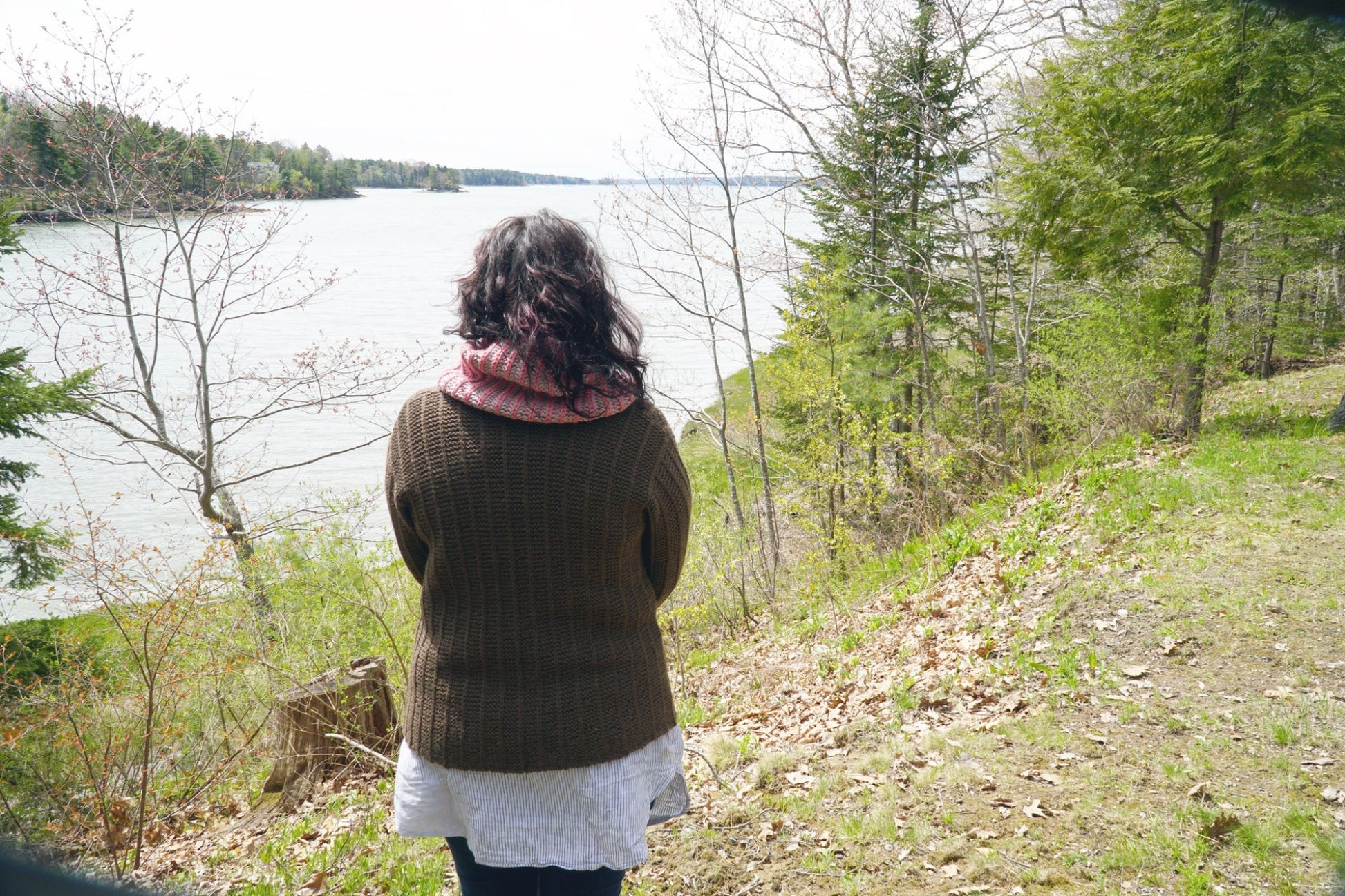 A person with long dark hair, wearing the Rangeley Cardigan by Halcyon Yarn in a Garter Rib stitch along with a white shirt and a red scarf, stands with their back to the camera, overlooking a lake surrounded by trees. The landscape is lush with greenery and the sky is overcast.