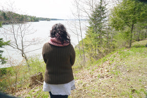 A person with long dark hair, wearing the Rangeley Cardigan by Halcyon Yarn in a Garter Rib stitch along with a white shirt and a red scarf, stands with their back to the camera, overlooking a lake surrounded by trees. The landscape is lush with greenery and the sky is overcast.