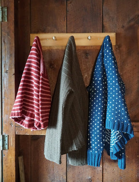 Three knitted sweaters hang on wooden pegs attached to a rustic wooden wall. The Halcyon Yarn Rangeley Cardigan is red with white stripes, the solid brown sweater is crafted from Norumbega yarn, and the blue one with white dots features a charming Garter Rib stitch pattern.