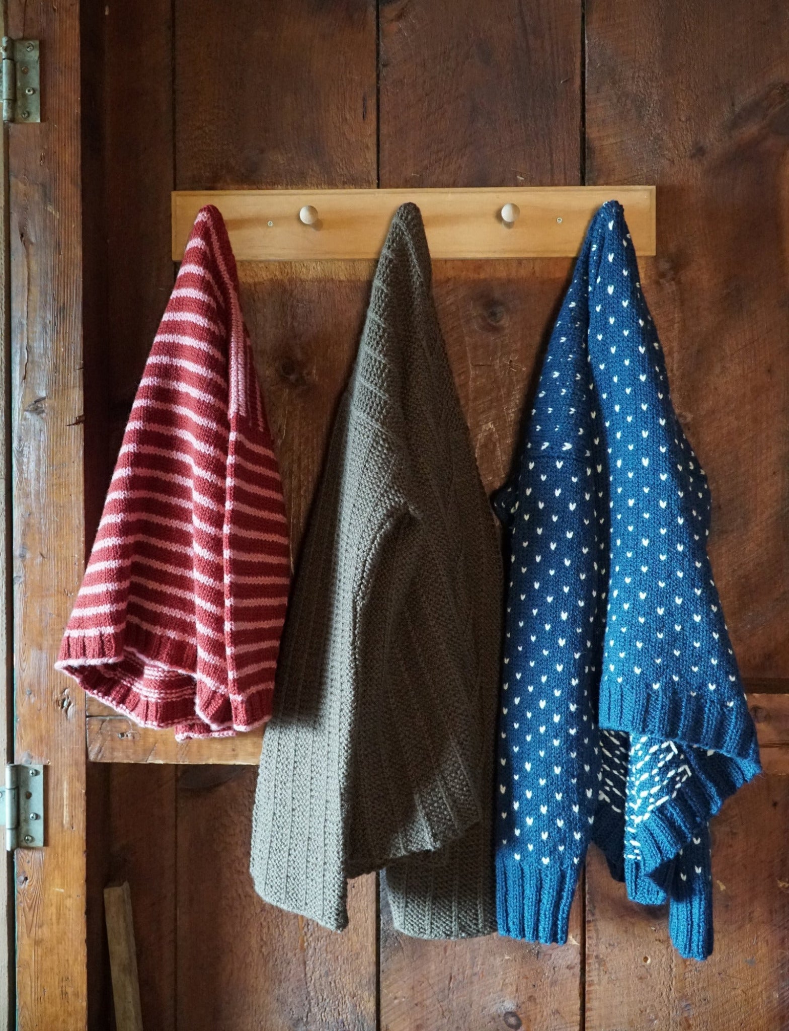 Three knitted sweaters hang on wooden pegs against a rustic wooden wall. From left to right: a red and white striped sweater, a solid brown Rangeley Cardigan from Halcyon Yarn, and a blue sweater with white polka dots. The setting gives a cozy, homely feel.