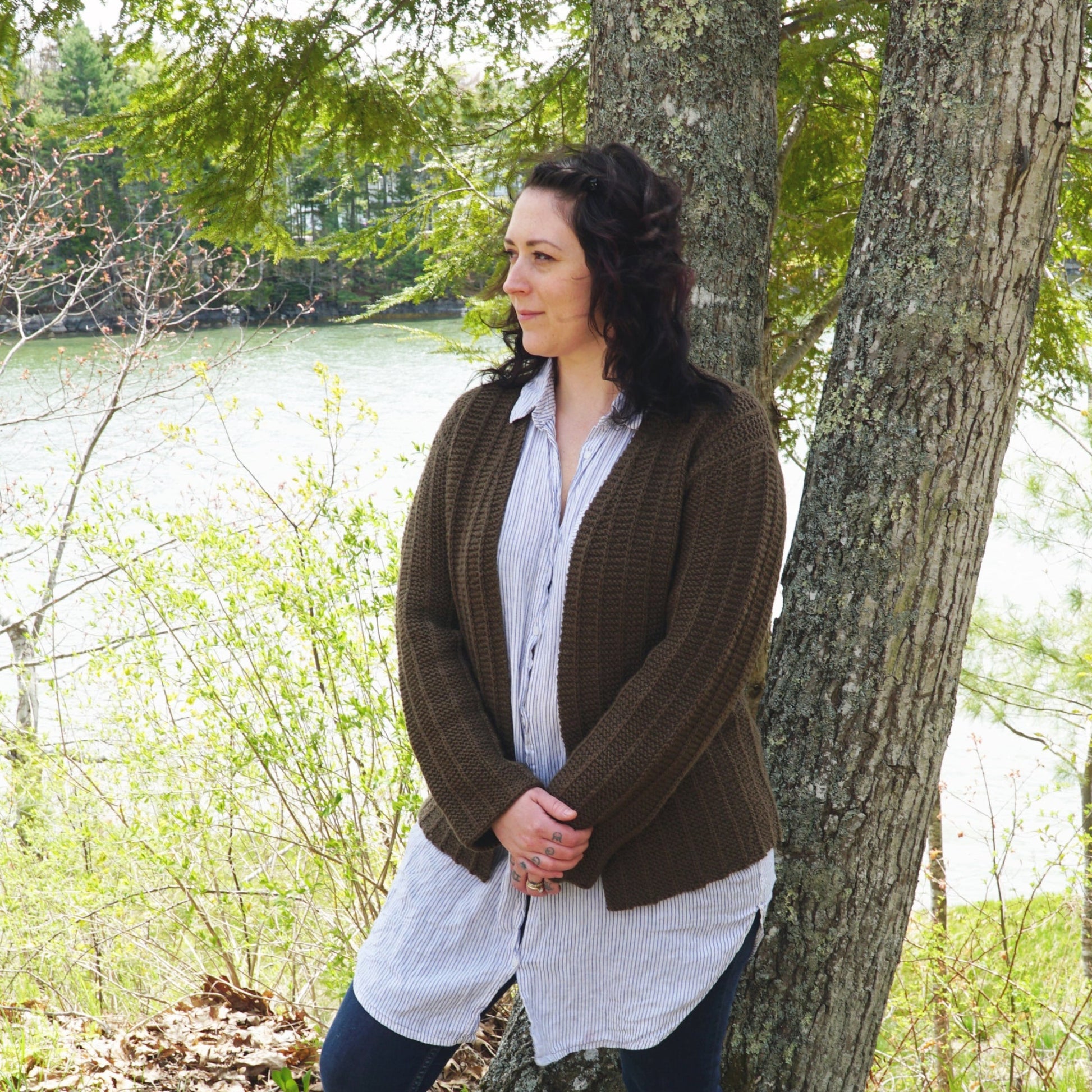 A person with curly dark hair, wearing a light striped shirt and the Halcyon Yarn Rangeley Cardigan made from Norumbega yarn in Garter Rib stitch, stands leaning against a tree trunk in a forested area. In the background, there is a body of water and greenery. They are looking off to the side with a serene expression.