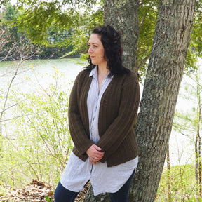 A person with dark, wavy hair stands leaning against a tree, looking to the side. They are wearing a long, striped shirt and a Rangeley Cardigan from Halcyon Yarn made from Norumbega yarn, paired with jeans. The background features a serene lake and lush green trees.