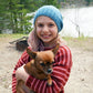 A young girl with wavy light hair, wearing a blue knit beanie and a red striped Lobster Roll Pullover by Halcyon Yarn in children's sizes, smiles while holding a small brown dog. They are outdoors near a body of water with trees in the background.